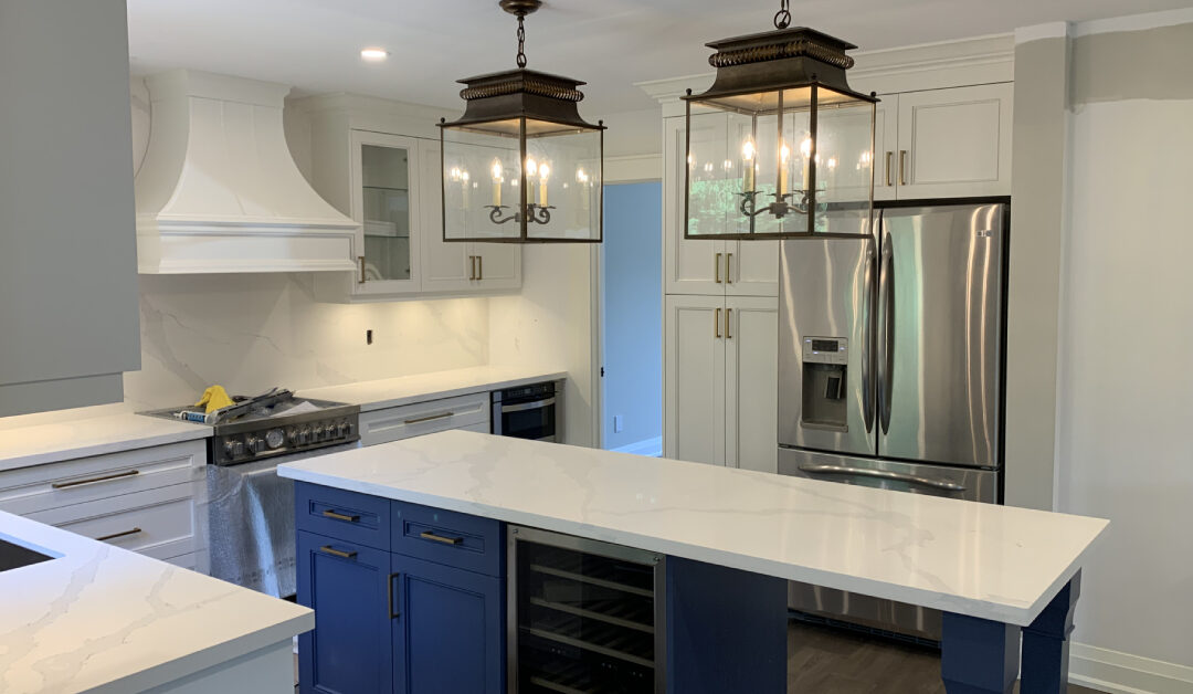 Timeless kitchen featuring white Shaker cabinets with gold hardware, a contrasting blue island with a veined white quartz countertop, stainless steel appliances, and two classic lantern pendant lights.