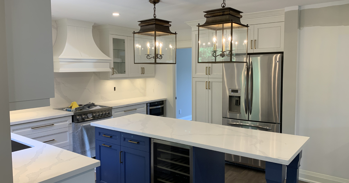 Timeless kitchen featuring white Shaker cabinets with gold hardware, a contrasting blue island with a veined white quartz countertop, stainless steel appliances, and two classic lantern pendant lights.