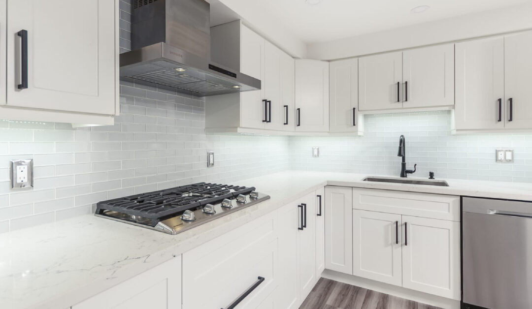 A modern kitchen featuring bright white custom shaker cabinets with matte black T-bar hardware, white quartz countertops with subtle gray veining, and a glossy light gray subway tile backsplash. Integrated under-cabinet LED lighting illuminates the countertops, highlighting a stainless steel gas cooktop, range hood, and a black gooseneck faucet, showcasing a completed custom cabinetry project in Burlington.