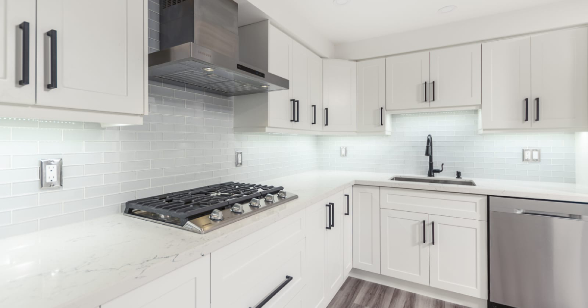 A modern kitchen featuring bright white custom shaker cabinets with matte black T-bar hardware, white quartz countertops with subtle gray veining, and a glossy light gray subway tile backsplash. Integrated under-cabinet LED lighting illuminates the countertops, highlighting a stainless steel gas cooktop, range hood, and a black gooseneck faucet, showcasing a completed custom cabinetry project in Burlington.