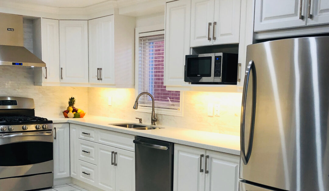 A bright, classic kitchen design featuring traditional white cabinets with brushed silver bar handles, clean white countertops, and a subtle stone tile backsplash. Warm under-cabinet lighting illuminates the counter space, which features a double-basin stainless steel sink and a gooseneck faucet. Modern stainless steel appliances, including a gas range, a chimney-style hood vent, a dishwasher, and a refrigerator, complete the functional layout, with a small microwave built into an upper cabinet shelf.