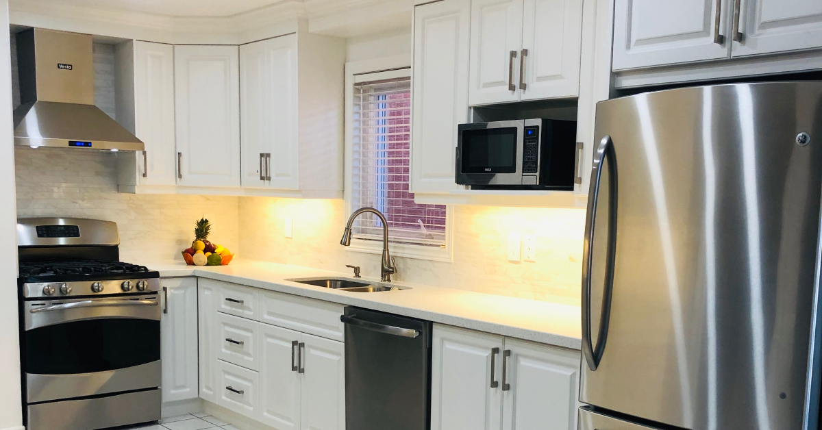 A bright, classic kitchen design featuring traditional white cabinets with brushed silver bar handles, clean white countertops, and a subtle stone tile backsplash. Warm under-cabinet lighting illuminates the counter space, which features a double-basin stainless steel sink and a gooseneck faucet. Modern stainless steel appliances, including a gas range, a chimney-style hood vent, a dishwasher, and a refrigerator, complete the functional layout, with a small microwave built into an upper cabinet shelf.