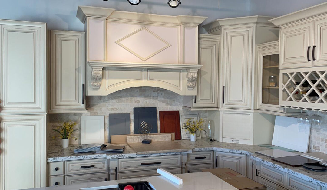 A traditional kitchen remodeling showroom display featuring cream-colored cabinetry with detailed molding, an ornate range hood, and a built-in wine rack. Various cabinet finish samples in navy, gray, and warm wood tones rest on the marbled quartz countertops, showcasing local design options for an Etobicoke kitchen renovation.