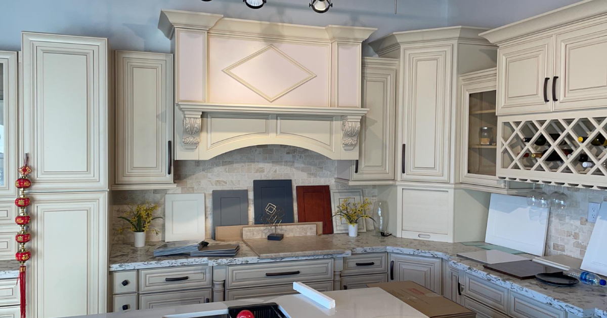 A traditional kitchen remodeling showroom display featuring cream-colored cabinetry with detailed molding, an ornate range hood, and a built-in wine rack. Various cabinet finish samples in navy, gray, and warm wood tones rest on the marbled quartz countertops, showcasing local design options for an Etobicoke kitchen renovation.