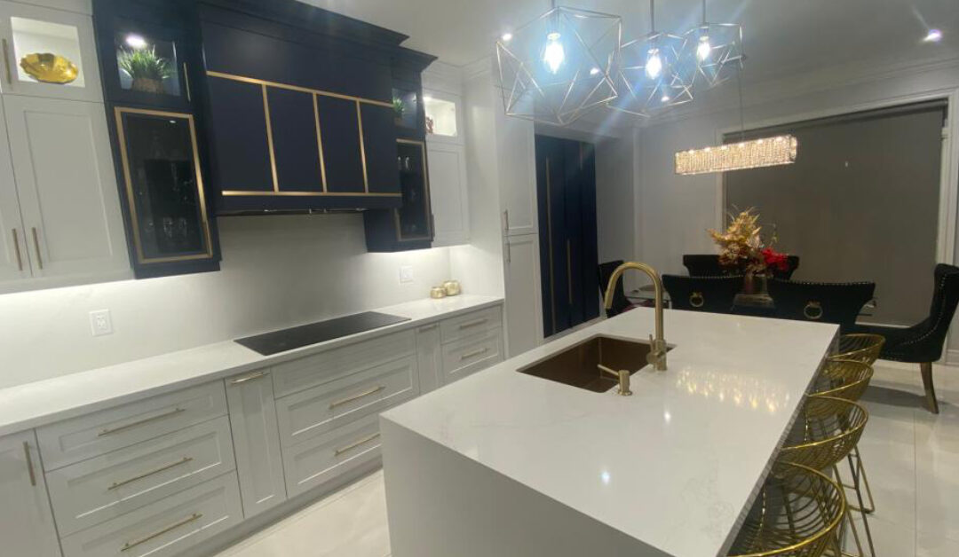 A luxurious modern kitchen upgrade featuring a high-contrast design with white shaker lower cabinetry and a dramatic navy blue custom range hood accented with gold geometric trim. A large white quartz waterfall island sits in the center, featuring an undermount gold sink, matching gold gooseneck faucet, and gold wire barstools. Geometric gold cage pendant lights hang overhead, beautifully tying together the brass cabinet hardware and elegant dining area in the background.