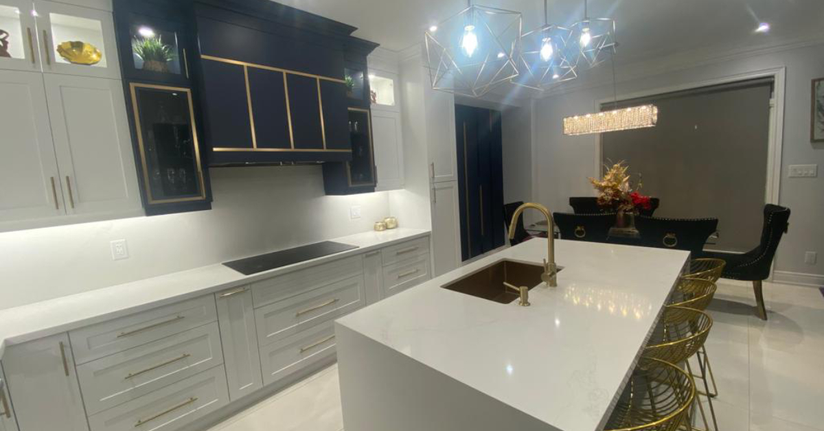 A luxurious modern kitchen upgrade featuring a high-contrast design with white shaker lower cabinetry and a dramatic navy blue custom range hood accented with gold geometric trim. A large white quartz waterfall island sits in the center, featuring an undermount gold sink, matching gold gooseneck faucet, and gold wire barstools. Geometric gold cage pendant lights hang overhead, beautifully tying together the brass cabinet hardware and elegant dining area in the background.