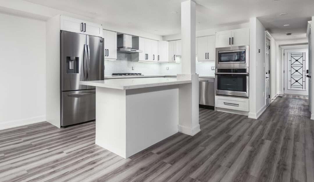 An open-concept luxury kitchen renovation featuring bright white shaker cabinets with slim black pull handles, white quartz countertops, and a central kitchen island integrated with a structural white pillar. Premium stainless steel appliances, including a double wall oven and a French-door refrigerator, contrast beautifully with the wide-plank gray wood-look flooring.