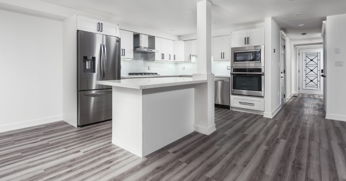 An open-concept luxury kitchen renovation featuring bright white shaker cabinets with slim black pull handles, white quartz countertops, and a central kitchen island integrated with a structural white pillar. Premium stainless steel appliances, including a double wall oven and a French-door refrigerator, contrast beautifully with the wide-plank gray wood-look flooring.