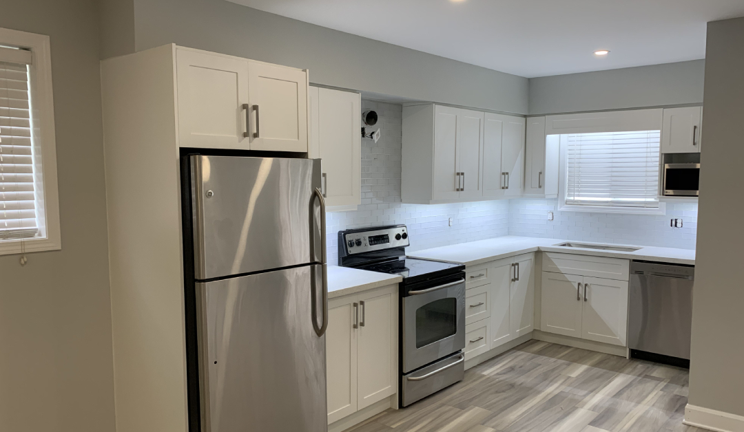 A modern L-shaped kitchen featuring white shaker cabinets, white countertops, and stainless steel appliances designed by Envyland Kitchen Cabinet.